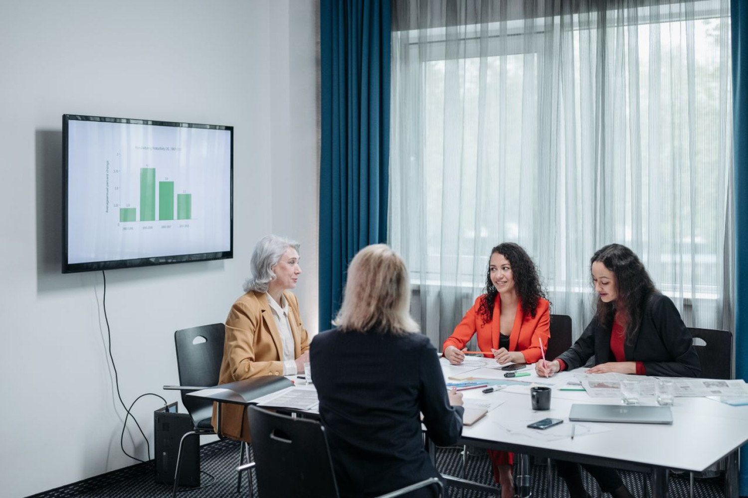 Vier Frauen besprechen an einem Tisch die Kosten eines LMS für Vereine, im Hintergrund zeigt ein Bildschirm ein Balkendiagramm